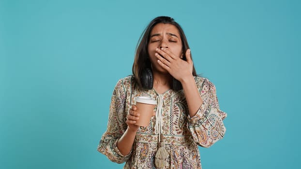 Woman suffering from insomnia yawning, sipping coffee to gain energy and get rid of pain. Tired person feeling drowsy after sleepless night, drinking caffeinated beverage, studio background, camera A