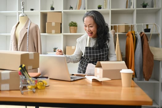 Happy senior woman small business owner looking at laptop screen and celebrating success.