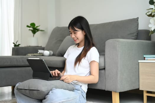 Happy young woman in casual clothes sitting on floor in living room and using digital tablet.