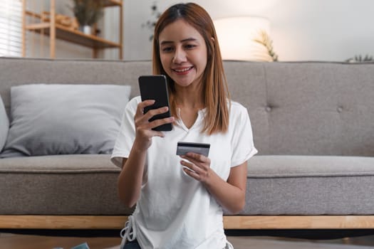 Young woman smiling while using a smartphone and holding a credit card, seated indoors on the floor