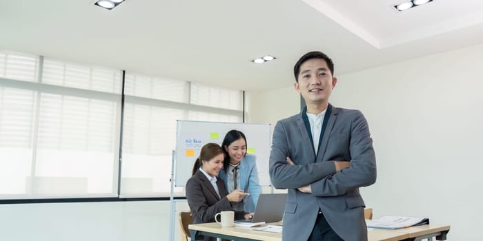 Confident business leader standing with arms crossed while his team collaborates on a laptop in the office, highlighting leadership and teamwork