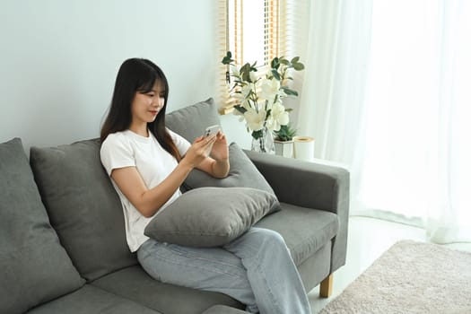 Asian female freelancer in casual clothes sitting on couch at home and using mobile phone.