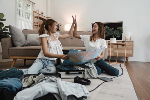 Two young women high-fiving while planning a trip with a map, surrounded by packed suitcases and clothing