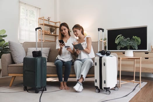 Two young women seated on a sofa, using a smartphone and credit card to book a hotel online, with suitcases nearby