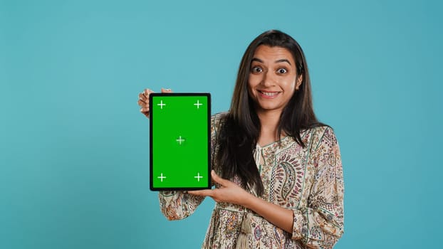 Portrait of indian woman doing influencer marketing using green screen tablet, isolated over studio background. Smiling person holding empty copy space mockup device, camera A