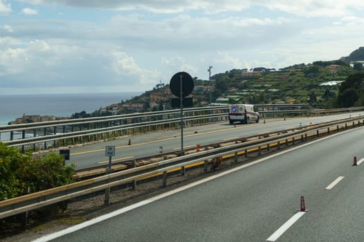 A van drives down a coastal highway in Italy, overlooking the Mediterranean Sea and a hillside town.