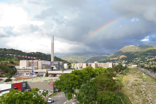 Mandelieu-la-Napoule, France - June 12, 2023: An industrial complex with a tall chimney is situated in a valley surrounded by mountains, with a rainbow visible in the cloudy sky after a rain shower.