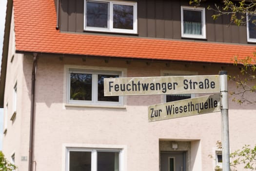 Two street signs point to Feuchtwanger Strabe and Zur Wiesehtquelle, in front of a white house with an orange roof.