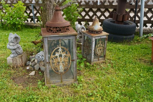 Two vintage, ornate lanterns sit amongst a lush green garden with a small, grey stone gnome statue and a large tree trunk nearby.