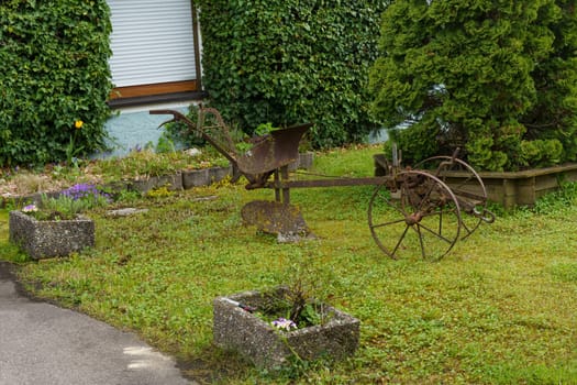 An old rusty farm implement is sitting on the grass in front of a house with green shrubbery.