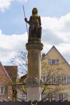 Feuchtwangen, Germany - June 6, 2023: A stone statue of a female figure holding a spear atop a fountain in a German town. The statue is decorated with intricate carvings.