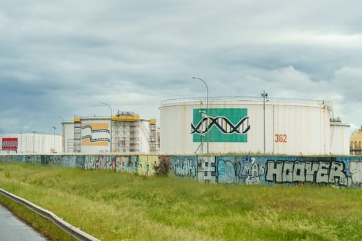 Lyon, France - May 7, 2023: Industrial oil tanks with murals and surrounding graffiti-covered wall are seen under a cloudy sky beside a grassy field.