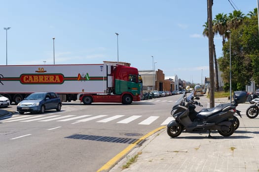 Barcelona, Spain - June 8, 2023: A large semi-trailer truck turns left at a street intersection in Spain. A scooter is parked on the sidewalk to the right of the image.