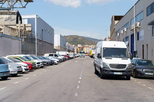 Barcelona, Spain - June 8, 2023: A white van is parked on the street in front of industrial buildings in Barcelona, Spain. Several cars are parked along the side of the street.