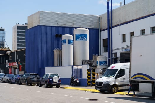 Barcelona, Spain - June 8, 2023: An industrial complex with large, cylindrical tanks labeled Linde and several delivery trucks parked in front. There are cars parked on the street nearby.