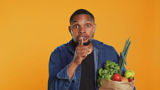 African american secretive guy doing mute hush gesture with finger over lips, keeping a secret about locally grown organic produce in a paper bag. Person being private about veganism. Camera A.