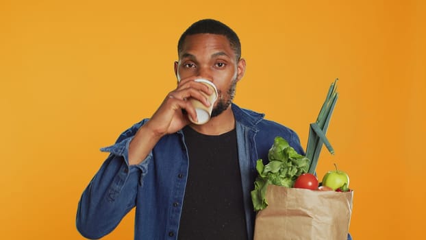 Relaxed carefree person drinking a coffee cup and carrying his groceries, going shopping at a local farmers market. Male model enjoying refreshment against orange background. Camera A.