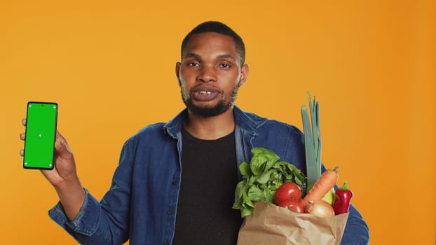 African american man showing a phone with isolated mockup screen, carrying a paper bag full of ethically sourced produce in studio. Model presenting greenscreen and eco friendly food. Camera A.