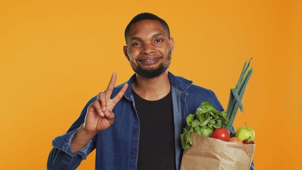 Young adult showing peace sign gesture and holding organic groceries, recommending sustainable lifestyle and homegrown produce. Peaceful relaxed guy carries eco friendly goods. Camera A.