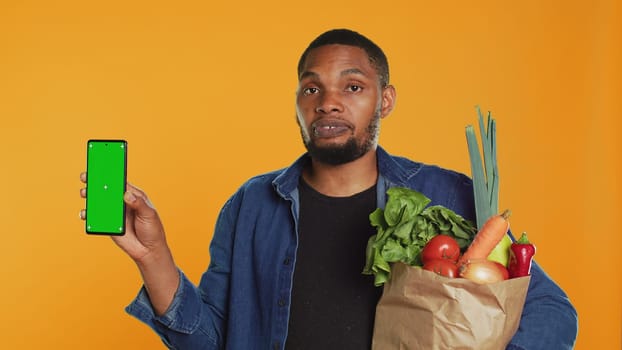 Young guy with a bag of fresh groceries showing greenscreen on his smartphone, presenting an isolated display on phone. Person advertising locally grown produce, zero waste. Camera A.