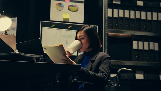 Private agent studying criminal files and archived folders in incident room, working on legal investigation to build suspects profile. Wheelchair user reading statements and reports. Camera B.