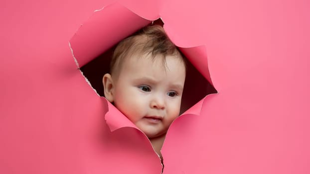 Cute Caucasian newborn baby boy peeks out of a hole in a paper pink background