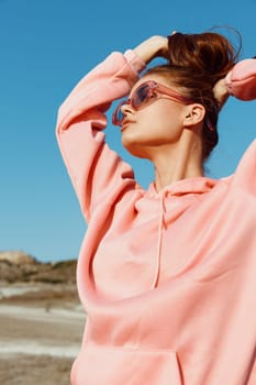 Stylish woman in pink sweatshirt and sunglasses standing on beach with hands on head enjoying the sun