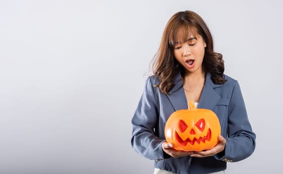 In this studio shot, a cheerful Asian woman cradles an orange pumpkin, radiating happiness, while surrounded by whimsical ghost pumpkins, setting the stage for a festive Halloween atmosphere.