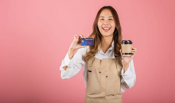 Portrait Asian happy young woman barista bar tender coffee maker holding coffee tea hot cup and credit card studio shot isolated on pink background, female smiling hold takeaways cup for payment