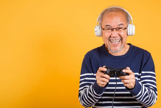 Portrait Asian smiling old man with headphones holding game controller studio shot isolated yellow background, senior man pensioner feeling happy playing a video game with joystick console