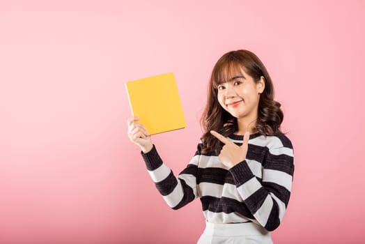 Portrait of happy Asian beautiful young woman confident smiling holding yellow book and pointing finger to the book, studio shot isolated on pink background, education concept