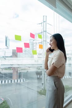 Businesswoman brainstorming ideas while looking at sticky notes on a window in a modern office setting, focusing on planning and creativity