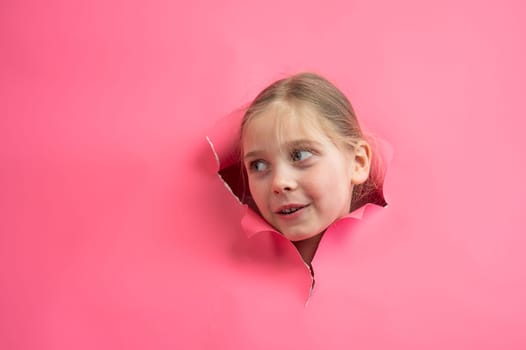 Cute Caucasian girl peeks out of a hole in a paper pink background