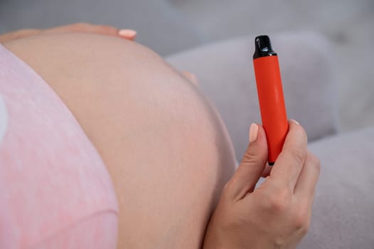 A pregnant woman smokes a vape while sitting on the couch. Close-up of the abdomen