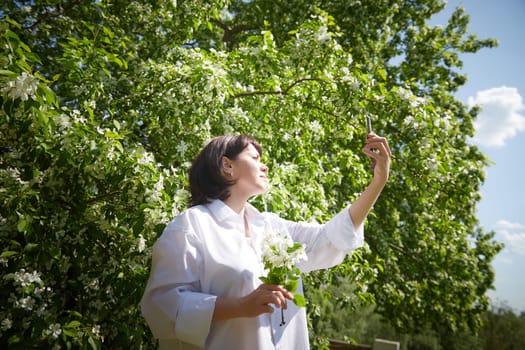 Brunette girl Using Smartphone in Blossoming Orchard at Springtime. Middle aged Woman taking selfie by phone among spring blossoms of apple trees