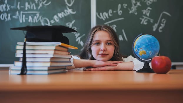 Adorable school girl posing at her desk against a background of blackboard, books, globe and graduation cap