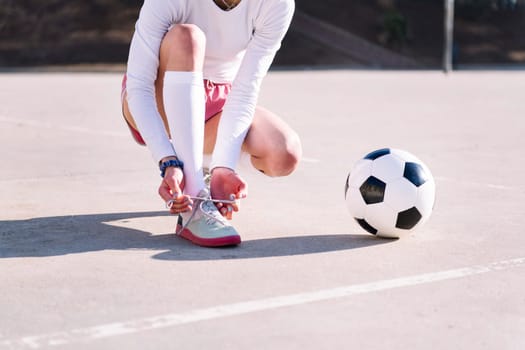 unrecognizable woman tying her sneakers next to a soccer ball to play in a urban football court, concept of sport and active lifestyle