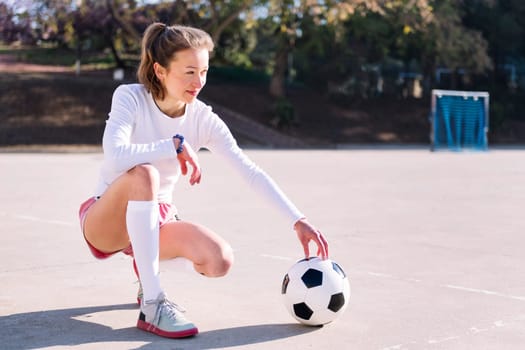 young caucasian woman squatting next to a soccer ball ready to play in a urban football court, concept of sport and active lifestyle