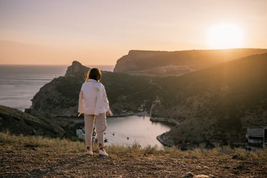 A woman stands on a hill overlooking a body of water. The sun is setting in the background, casting a warm glow over the scene. The woman is enjoying the view and taking in the beauty of the landscape