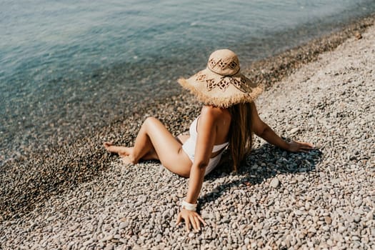 Beach Relaxation woman in hat sits on a pebble beach enjoying the sunshine. The concept of travel, vacation at sea.