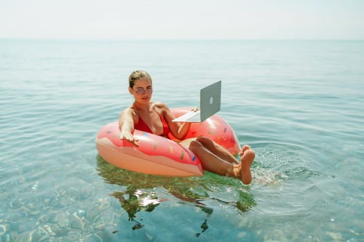 Woman laptop sea. Freelancer woman in sunglases floating on an inflatable big pink donut with a laptop in the sea. People summer vacation rest lifestyle concept