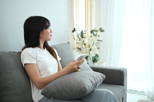 Attractive asian young woman relaxing on couch at home holding mobile phone and looking outside window.