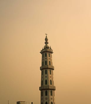 The image captures a tall, intricately decorated minaret of a mosque against a soft, golden sunset sky. The minaret is adorned with intricate details, including multiple tiers and arched windows. It stands tall and slender, reaching towards the sky. The background sky is a muted, warm hue, creating a peaceful and serene atmosphere.