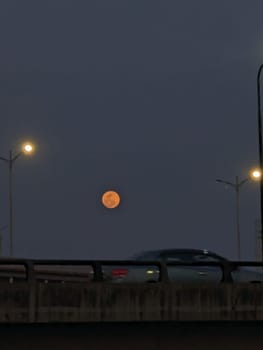 This image shows a full moon rising in the sky over a city street at dusk. The moon is a bright orange color and appears to be full. Streetlights illuminate the scene, casting a warm glow. A car is driving on the road below, which is visible as a dark silhouette against the bright moon.