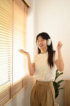 Happy young asian woman holding phone listening to favorite songs and dancing in living room.