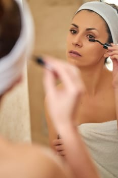 Beautiful young brunette woman applying mascara on eyelashes in front of bathroom mirror