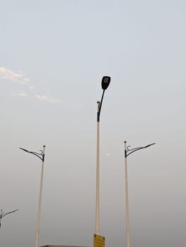 This image shows three streetlights against a hazy, pale blue sky at dusk. The streetlights are tall and slender, with white poles and black arms that extend out to the side. The streetlights are casting long shadows on the ground below. The sky is mostly clear, with just a few wisps of clouds visible. The overall mood of the image is peaceful and serene.