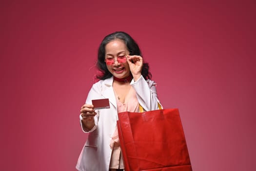 Portrait happy mature woman holding shopping bag and credit card over red background.