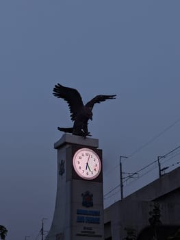 The image showcases a clock tower in the Philippines, featuring a prominent eagle statue perched atop. The clock face glows brightly against the dusky sky, illuminating the time. The surrounding area features a concrete structure and overhead power lines, hinting at an urban setting.