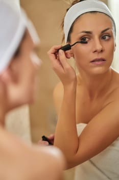 Beautiful young brunette woman applying mascara on eyelashes in front of bathroom mirror